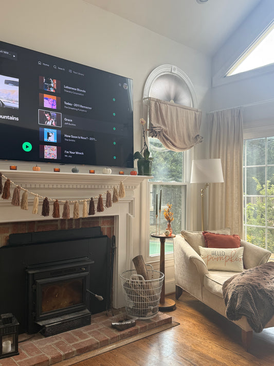 Living room with a large TV screen displaying a music app, fireplace, and cozy fall decor including tassel garland, colorful pumpkins, and small votives.