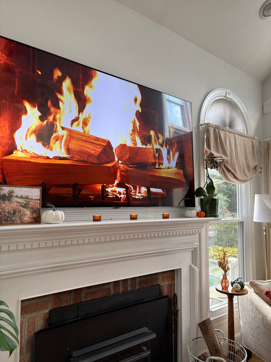 Mantel in a living room decorated with a fall painting, pumpkins, and votives with a large TV displaying a fireplace.
