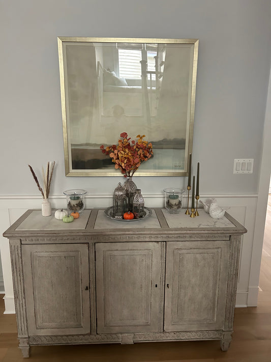 Decorative sideboard with pumpkins, vase, and candlesticks with a painting above it in a living room.