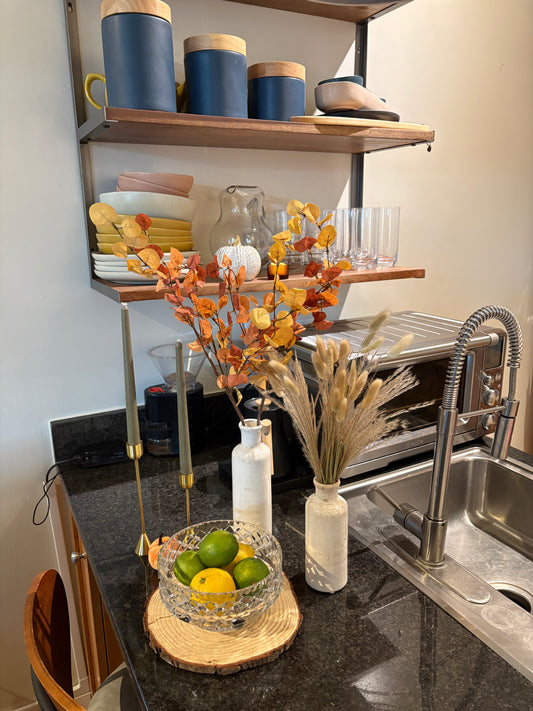Fall kitchen decoration with candlesticks, vases, pampass grass, and wooden round with glass bowl of fruit on top.
