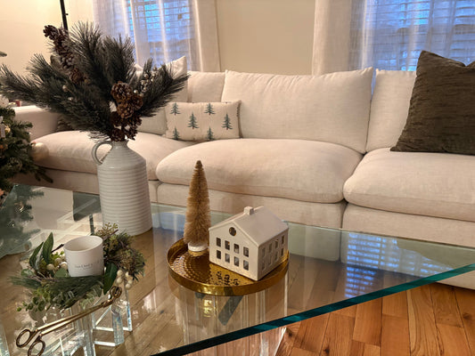 Neutral winter decor vignette on a coffee table in a living room with natural greenery and pinecones in a vase, gold tray with bottle brush tree and ceramic house, and candle with greenery candle ring.