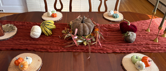 Thanksgiving table set with rust table runner, woven brown pumpkin centerpiece in leaf wreath with candlesticks and gourds on the sides. Wooden tree rounds serve as chargers with pumpkins on top.