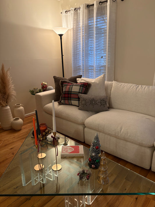Living room with a beige sofa, glass coffee table, and decorative Christmas items including pillows, candles, trees.