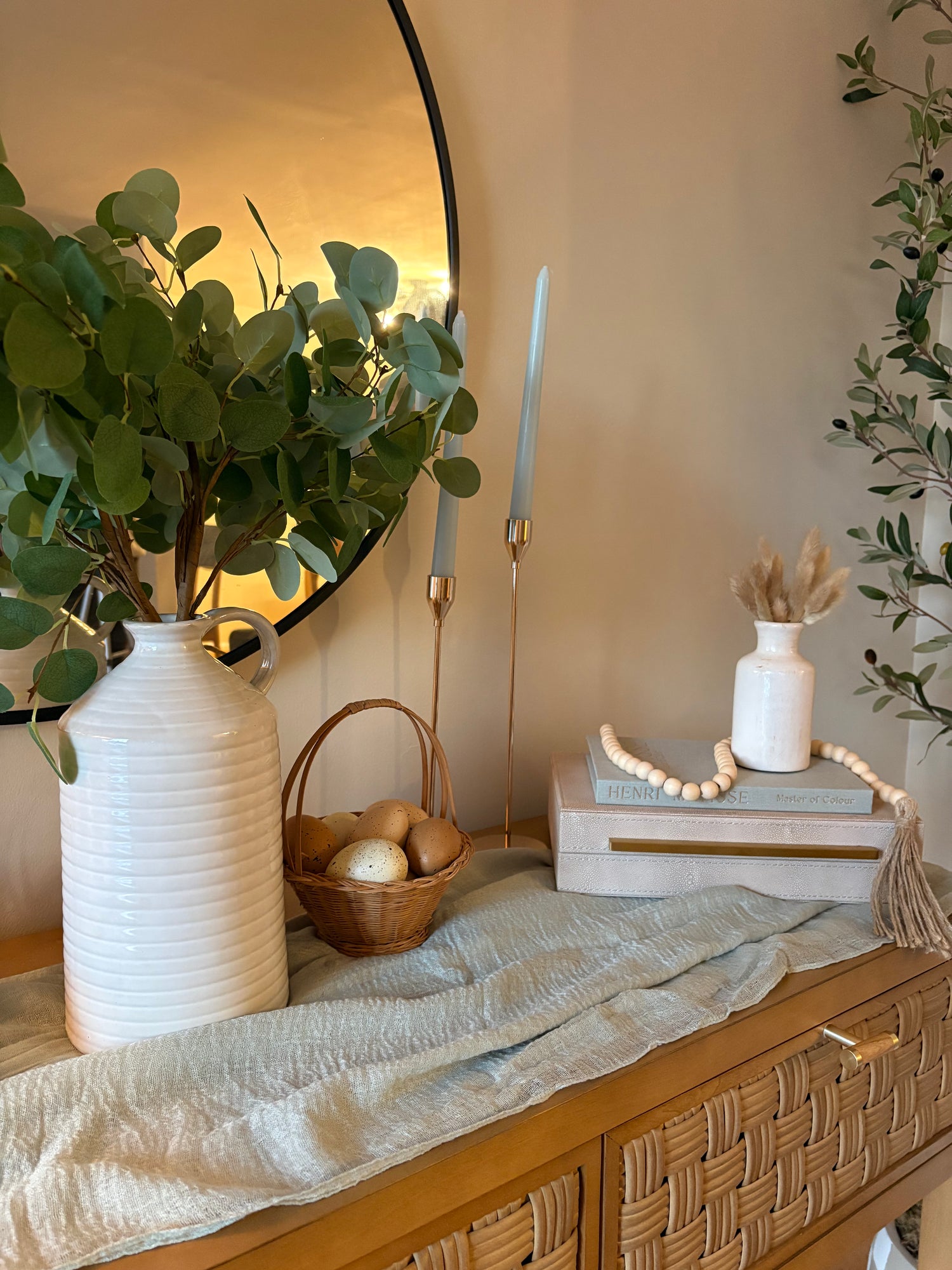 Entryway console table decorated with spring rental decor including a light green runner, a cream vase filled with faux eucalyptus, gold candlesticks with light blue tapered candles, and a box with book stacked on top of it with a beaded garland and small vase and faux filler.