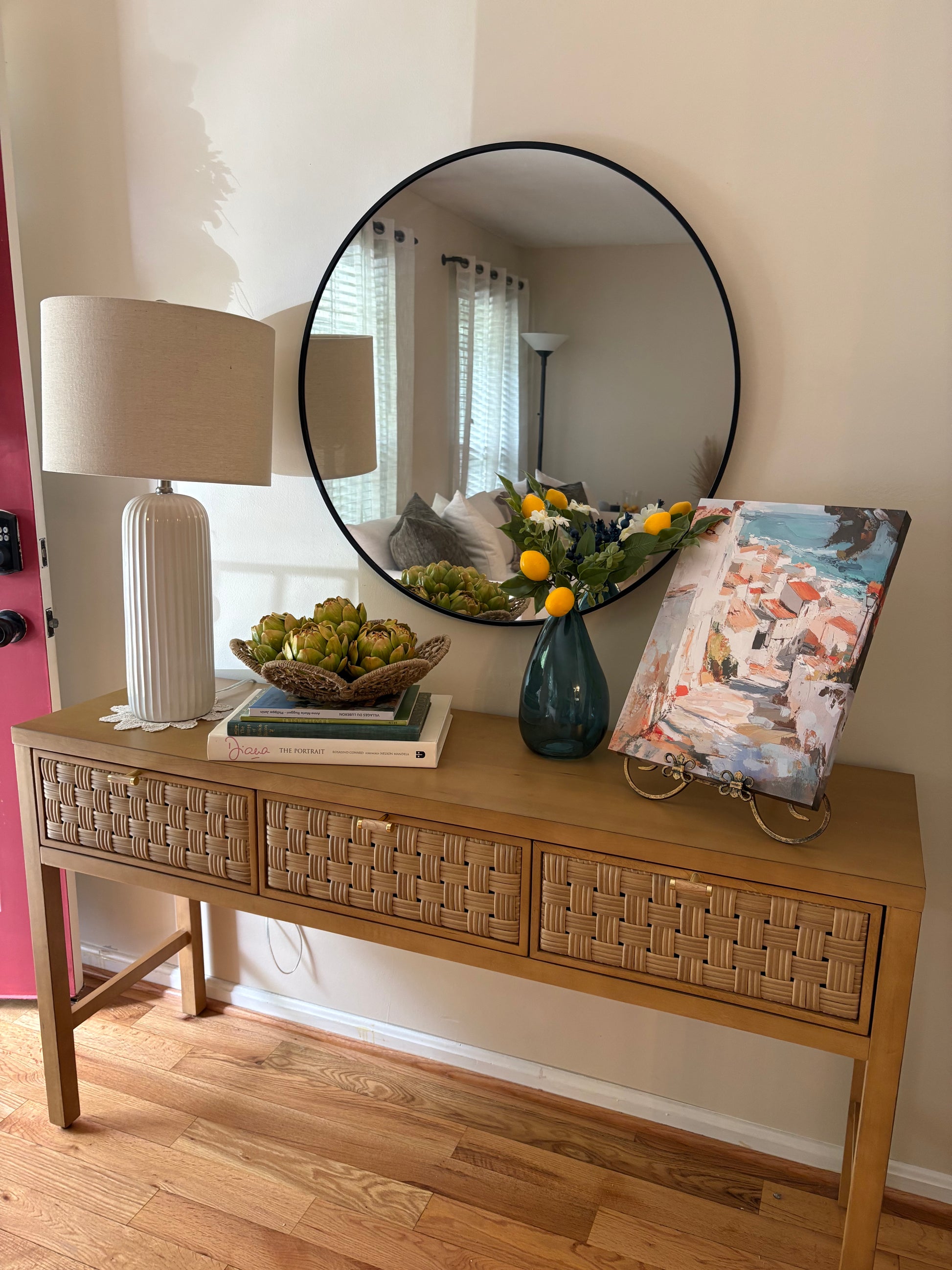 Wooden console table with summer rental decor including stack of books, faux artichokes in a bowl, blue vase with lemon and greenery filler, and a painting of a beachside villa.