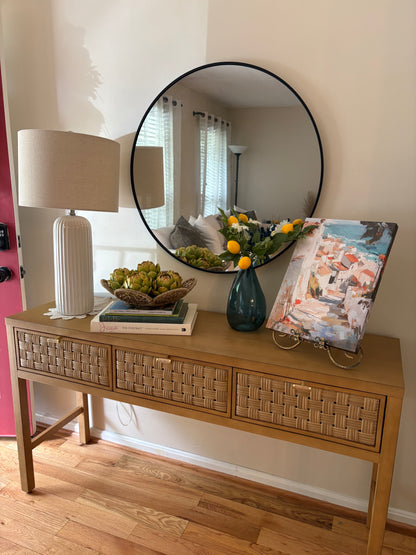 Wooden console table with summer rental decor including stack of books, faux artichokes in a bowl, blue vase with lemon and greenery filler, and a painting of a beachside villa.