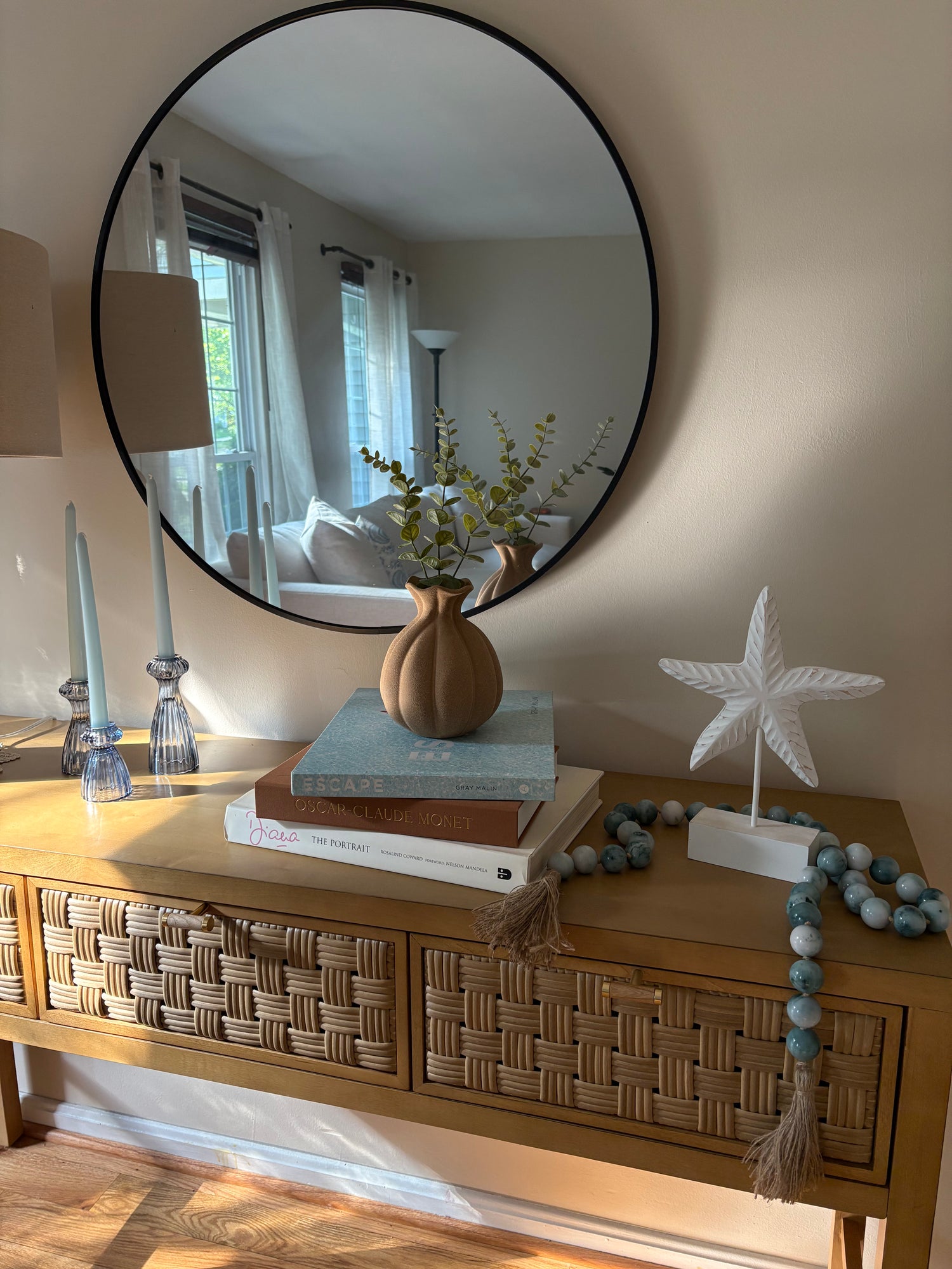 Wooden console table with nautical themed summer rental decor including glass candlesticks and tapered candles, a stack of decorative books, vase with eucalyptus, white starfish sculpture, and beaded tassel garland.