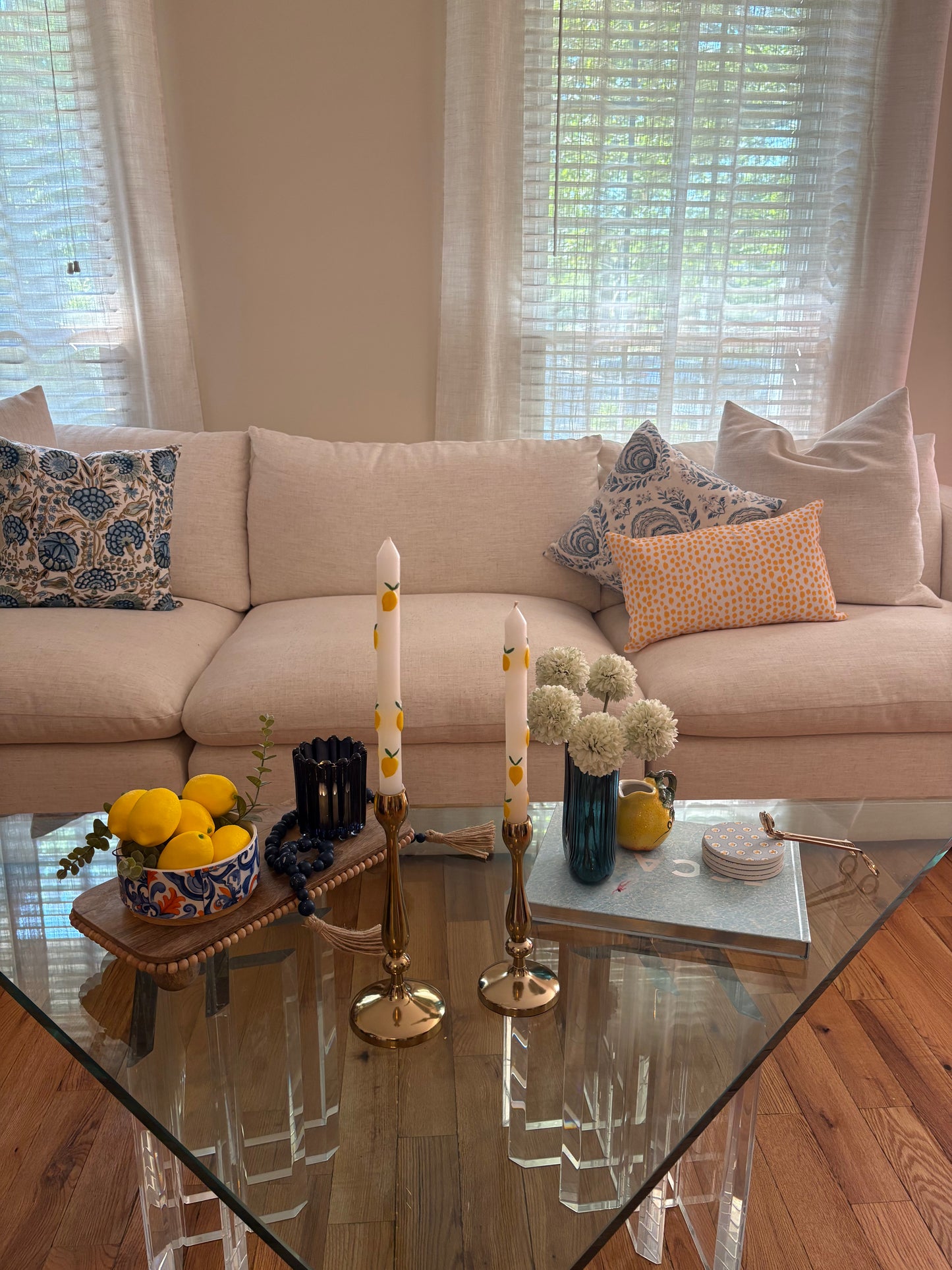 Living room with beige sofa and glass coffee table styled with summer rental decor including wooden tray with bowl of lemons, blue beaded tassel garland, candlesticks with tapered candles, decorative book stacked with coasters, vase with faux flowers, and wick trimmer.