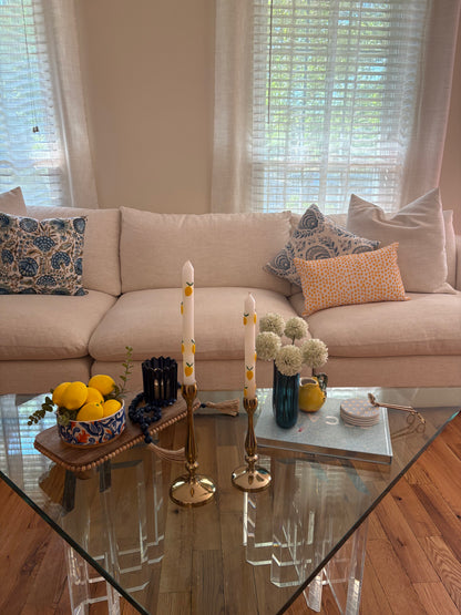 Living room with beige sofa and glass coffee table styled with summer rental decor including wooden tray with bowl of lemons, blue beaded tassel garland, candlesticks with tapered candles, decorative book stacked with coasters, vase with faux flowers, and wick trimmer.