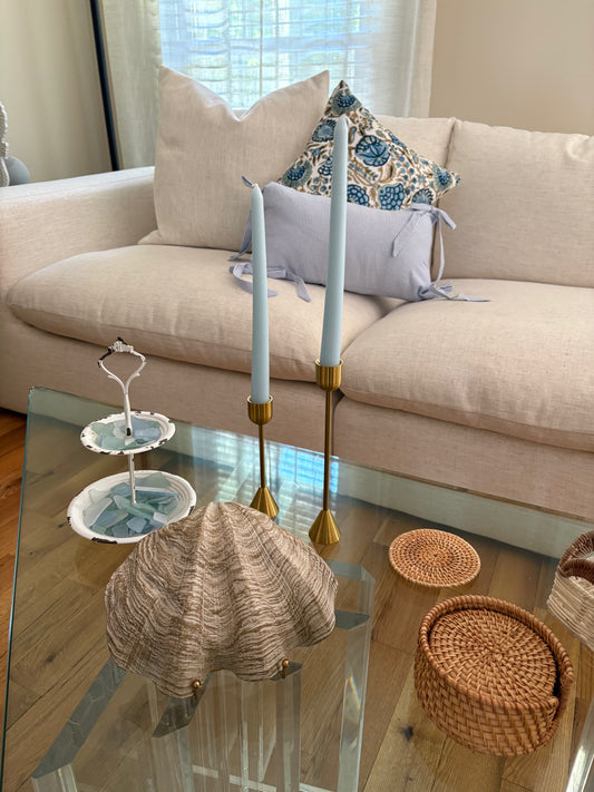 Living room with a beige sofa and glass coffee table with summer rental decor including sea glass, seashell, candlesticks with blue tapered candles, rattan coasters, and blue throw pillows.