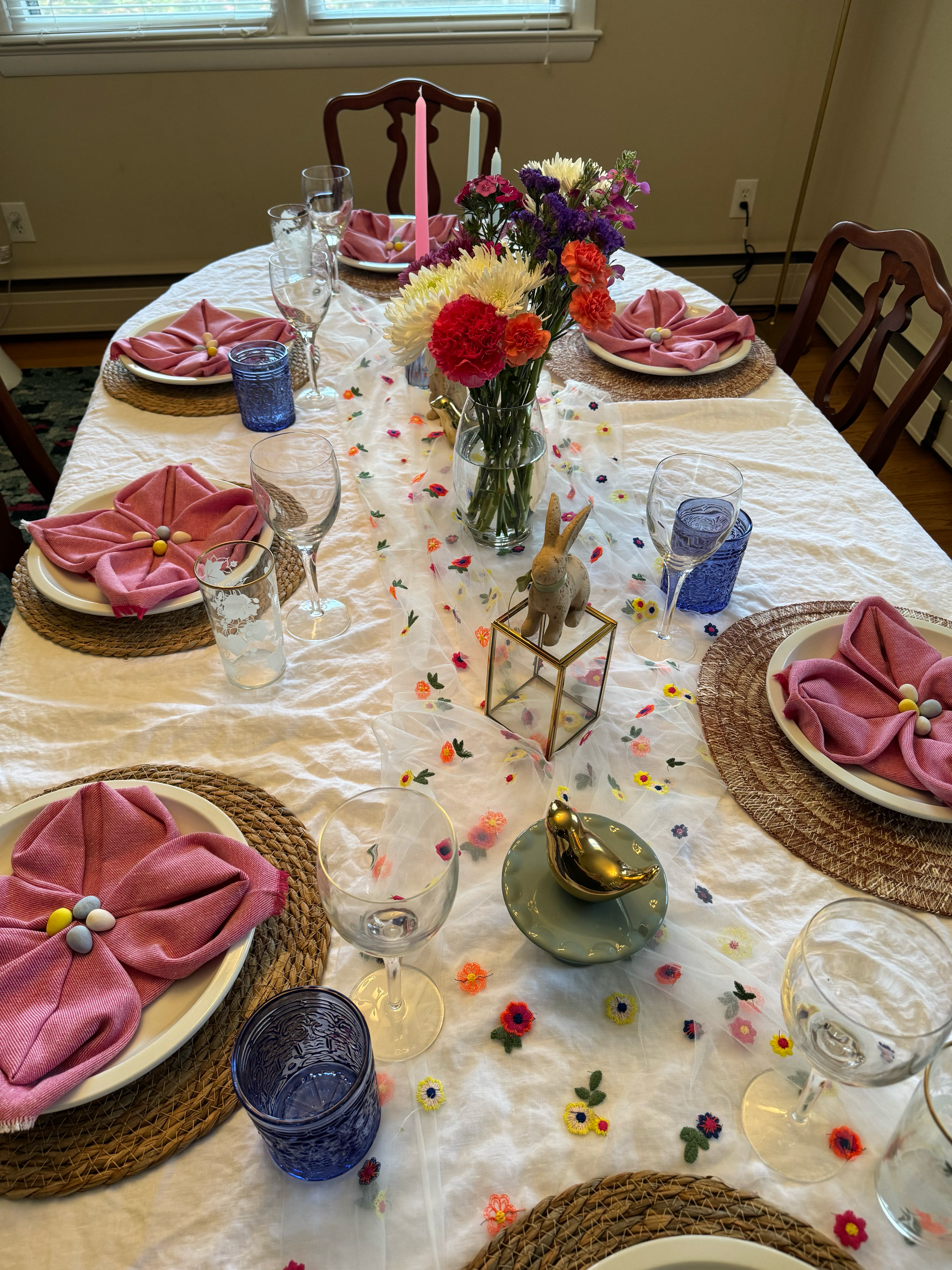 Dining table decorated for Easter with rental decor including vases, table runner with flowers, pink napkins, bird and bunny accent pieces, and colorful glasses.