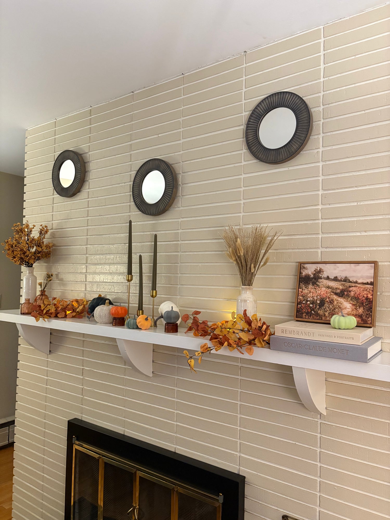 Decorative fireplace mantel with fall-themed decor including pumpkins, faux flowers, vases, and decorative books against a tiled wall.