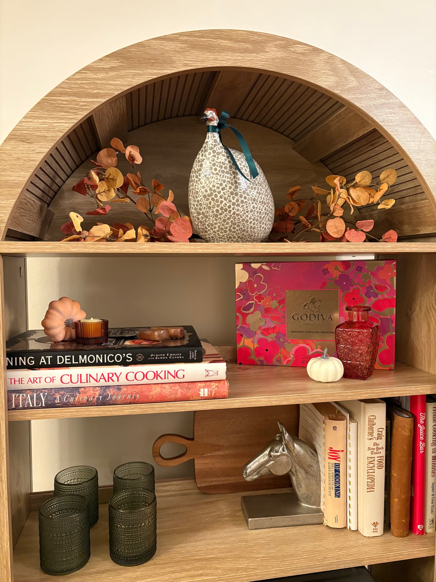 Wooden bookshelf with decorative items including books, a vase, pumpkins, fall florals, and a Godiva chocolate box.