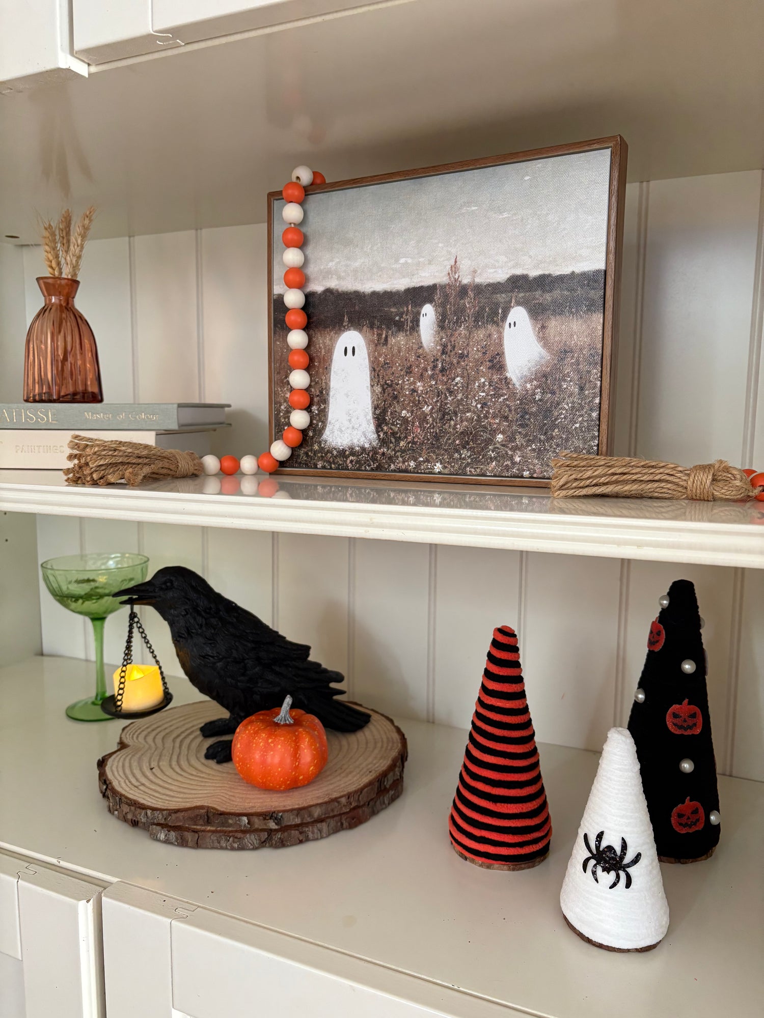 Halloween-themed decor on a shelf with a framed picture of ghosts, a crow, pumpkin, and decorative cones.