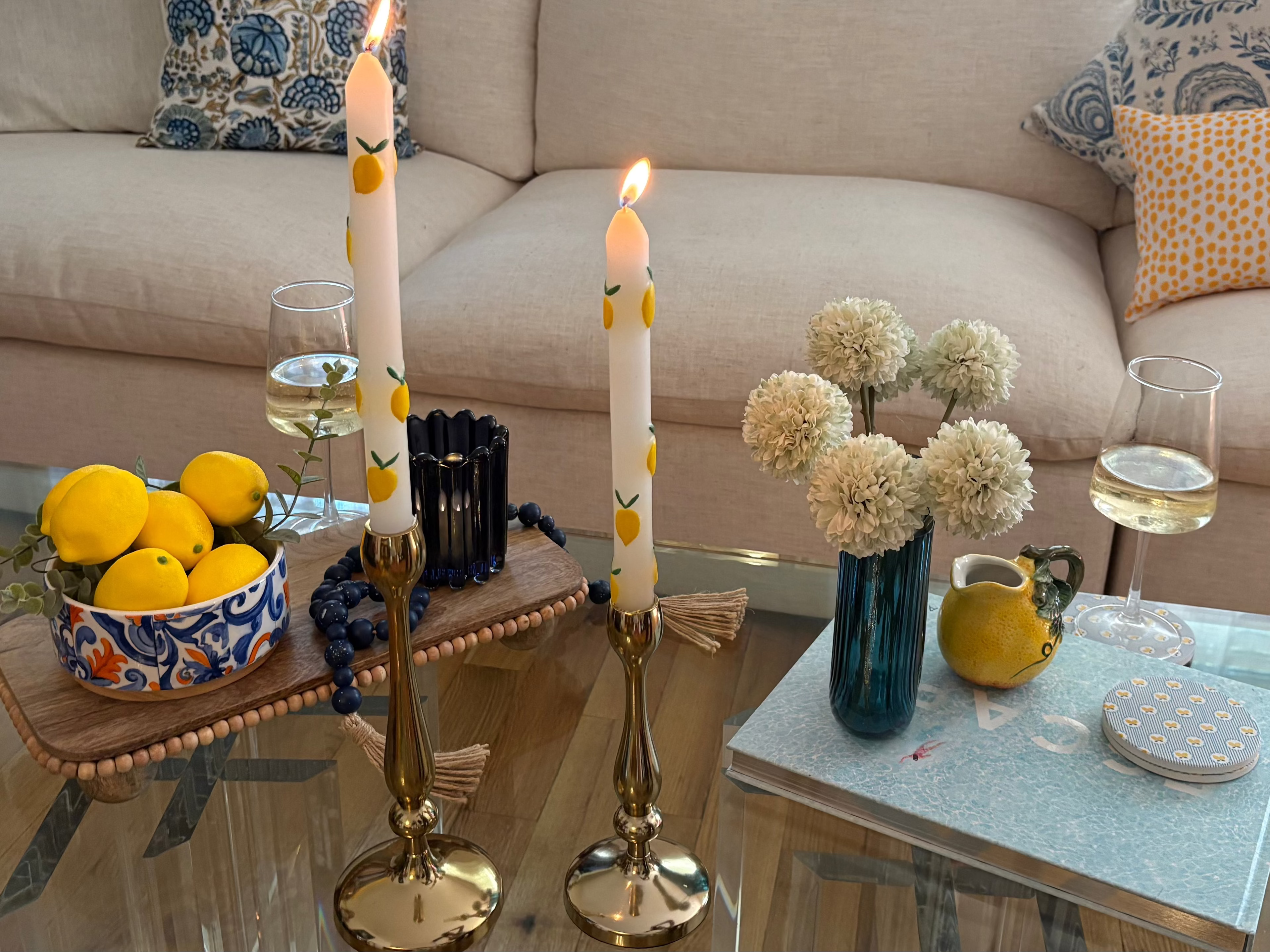 Living room with a beige sofa, glass coffee table with summer rental decor including wooden tray, bowl with lemons, blue glass candle holder, tapered candles with lemons, decorative book, vase with faux flowers