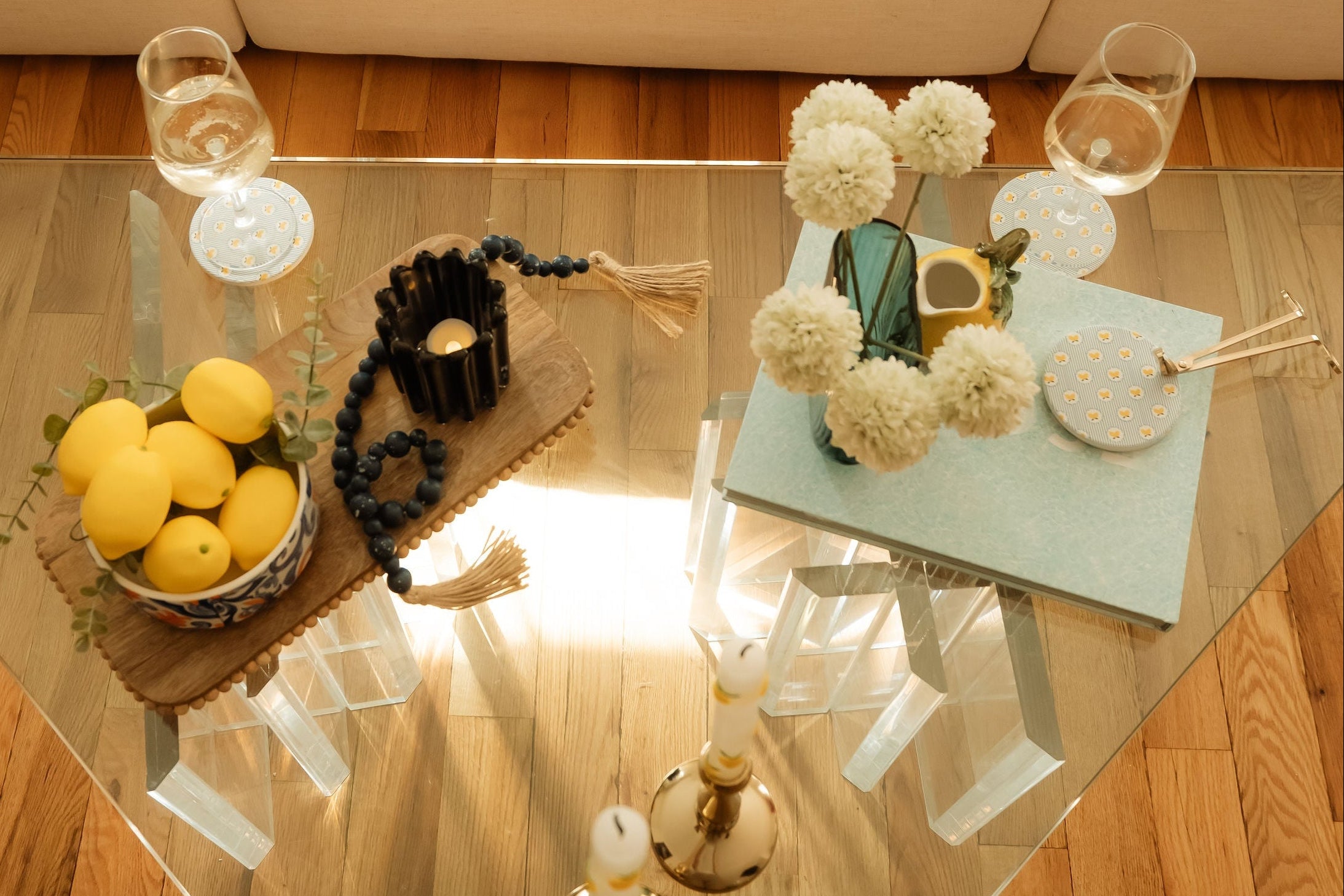 Glass coffee table with summer rental decor including decorative books, coasters, vase, faux flowers, lemons, beaded garland, and candles in a living room setting