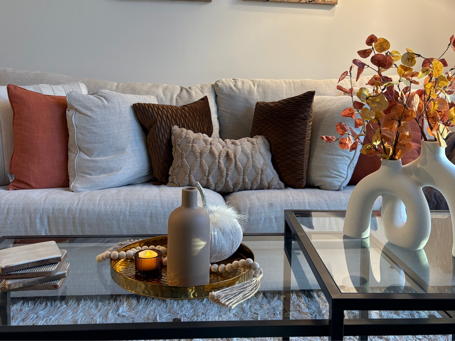 Living room decorated for fall with a couch, coffee table, and decorative elements including a gold tray, brown vase, beaded garland, small tea light, white vase, and orange fall vase filler.