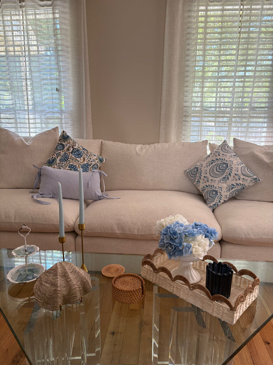Living room with beige sofa and nantucket themed summer rental decor including blue throw pillows, and a glass coffee table with sea shell, rattan coasters, candlesticks with tapered candles, rattan tray with hydrangeas and blue glass candle holder.