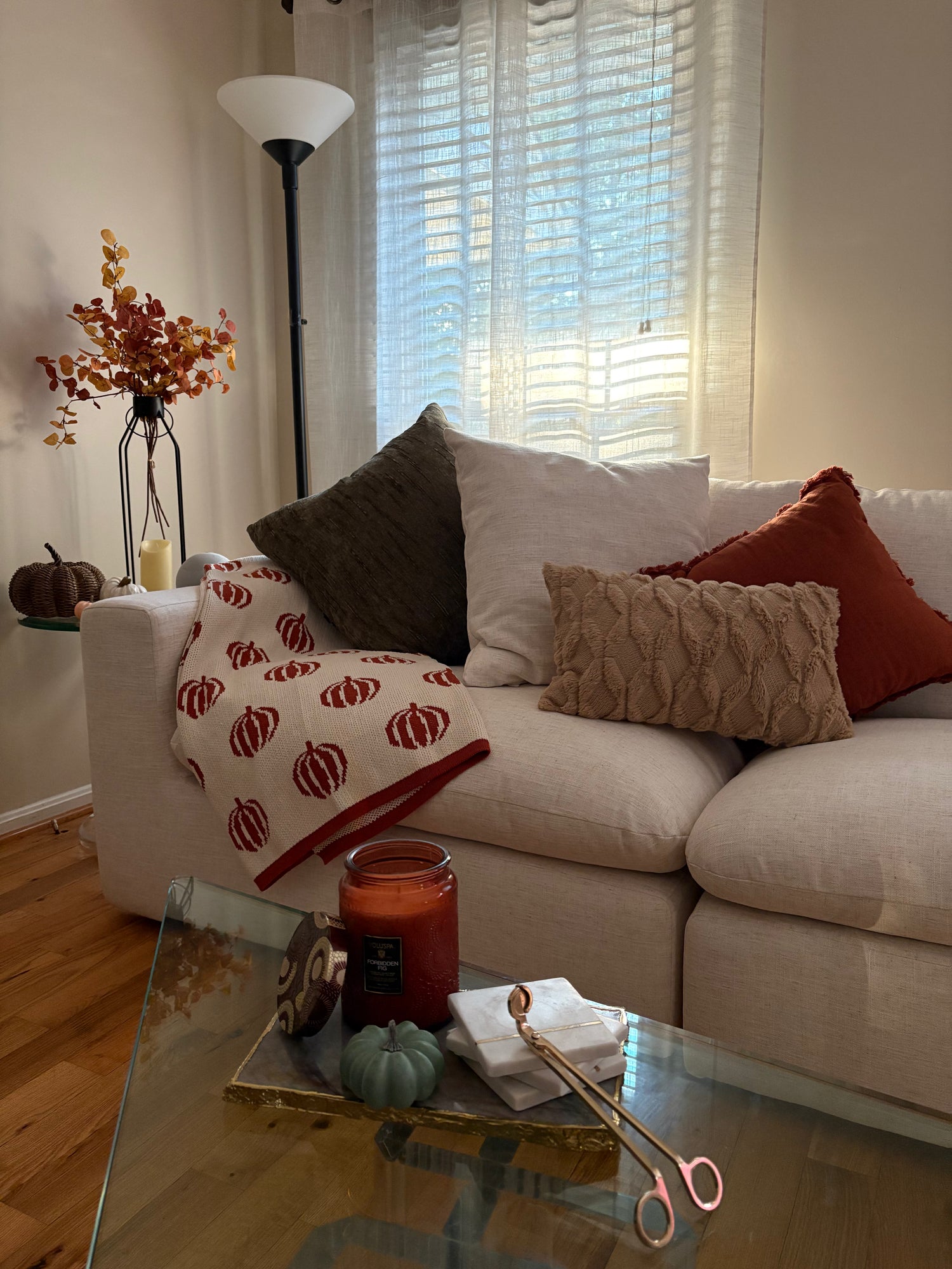 Living room with a beige sofa adorned with decorative pillows, a glass coffee table with candles and decor items, and a vase with autumn leaves.
