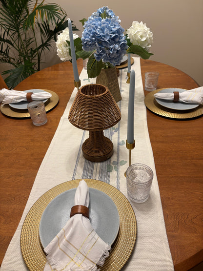 Elegant summer dining table setting with rental decor including floral centerpiece, candlesticks with tapered candles, rattan lamp, white and blue table runner, and decorative plates.