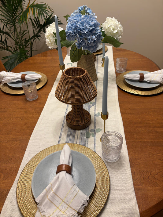 Elegant summer dining table setting with rental decor including floral centerpiece, candlesticks with tapered candles, rattan lamp, white and blue table runner, and decorative plates.