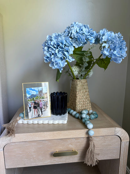 Decorative table with summer rental decor including blue hydrangeas, a photo frame, marble scalloped tray, candles, and beads.