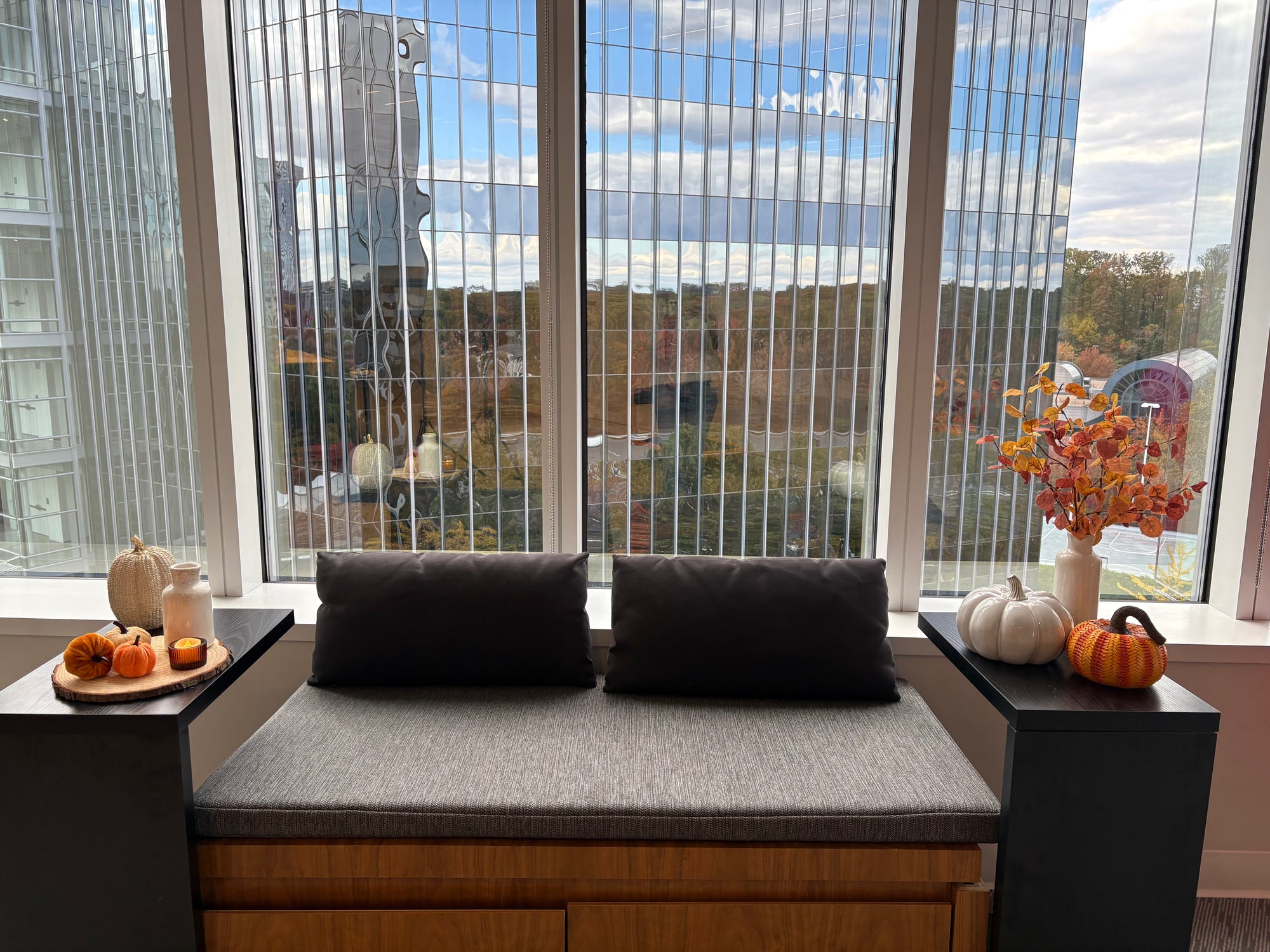 Window seat couch with pillows and fall decorative items including wooden stump slice with pumpkins and vases on both sides in a room with large windows.