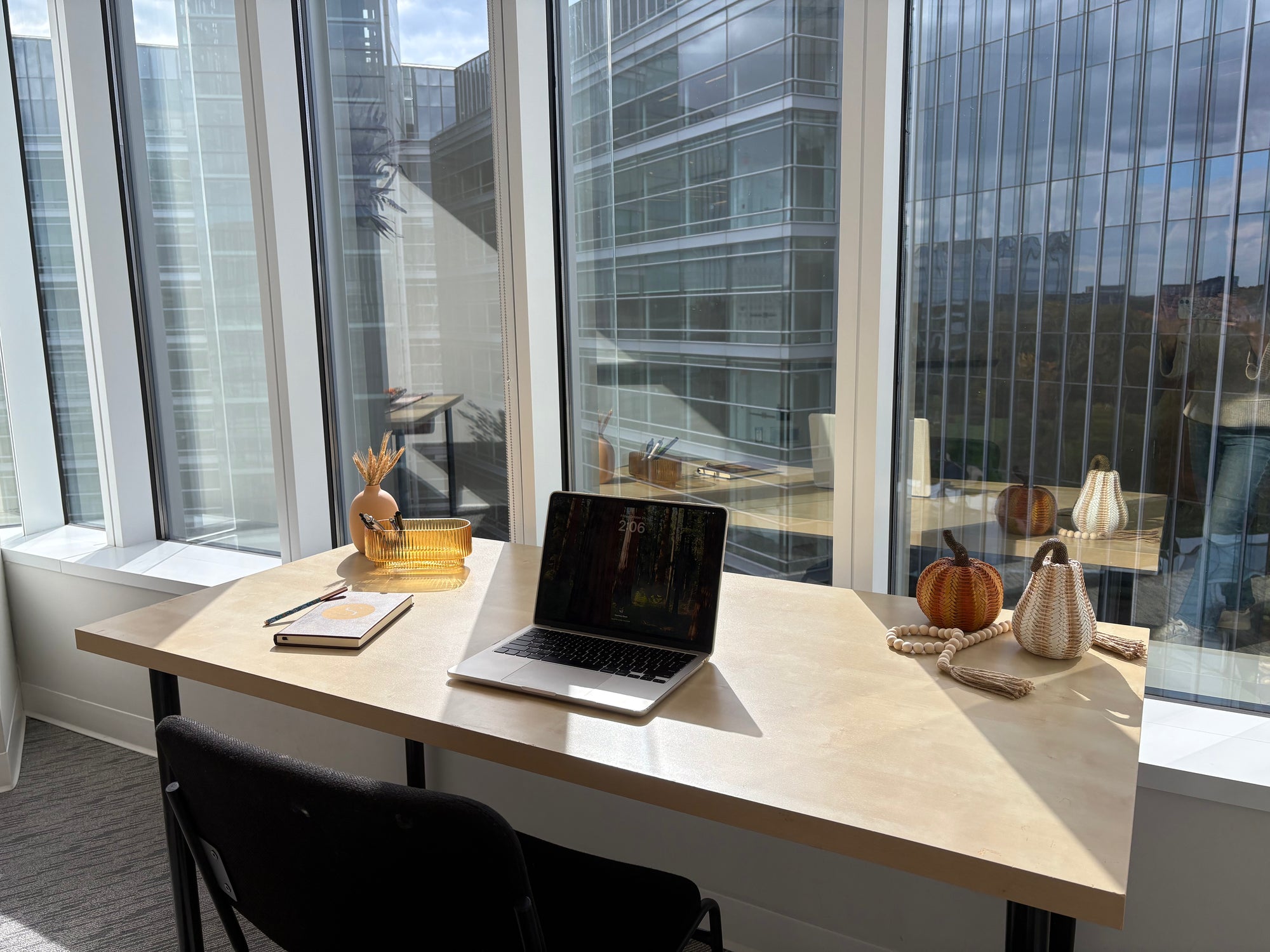Modern office desk with laptop, decor items including textured pumpkins and gourds, a vase with filler, and a glass ribbed pencil holder in front of large windows.