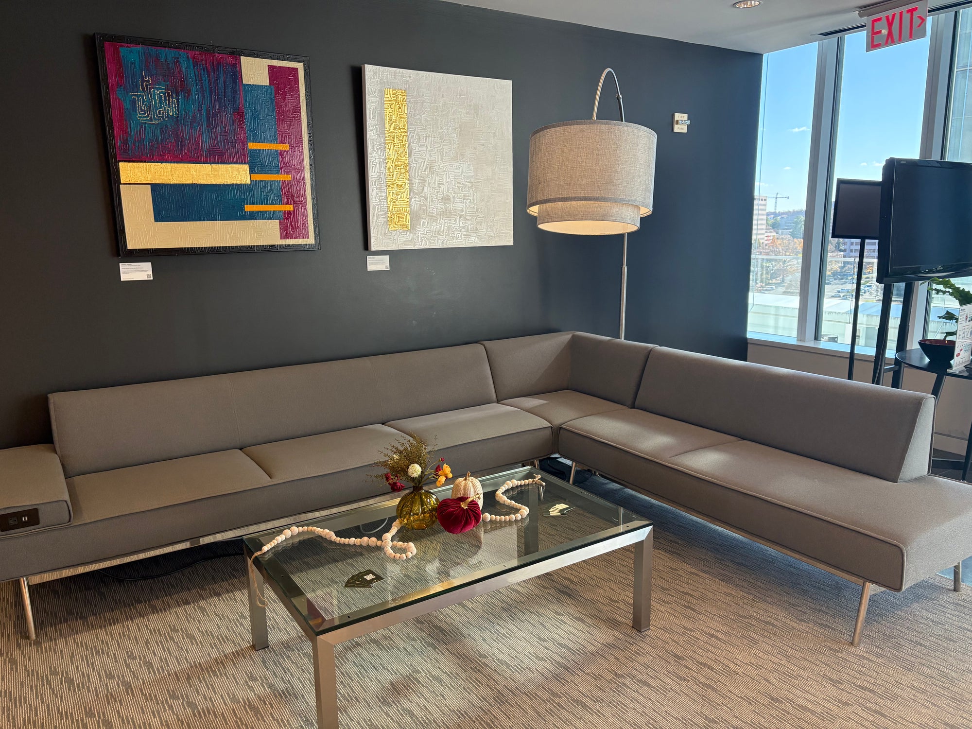 Modern office sitting room area with a sectional sofa, glass coffee table with fall decorations including velvet pumpkin, ceramic white and gold pumpkin, and vase full of flowers and greenery with abstract art on the wall.
