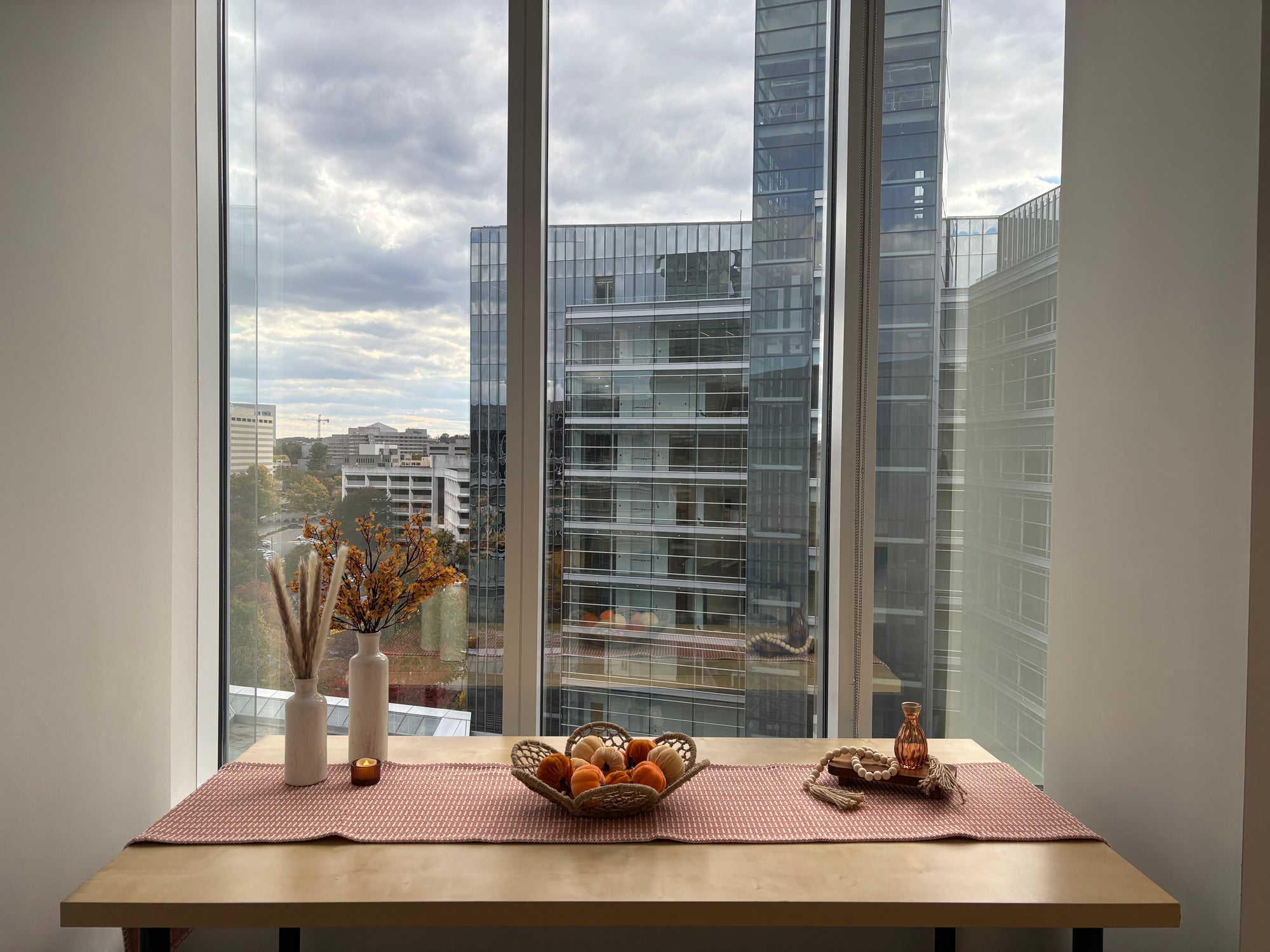 Table with decorative fall items including vases, floral vase filler, basket of pumpkins, and table runner front of a large window with cityscape view