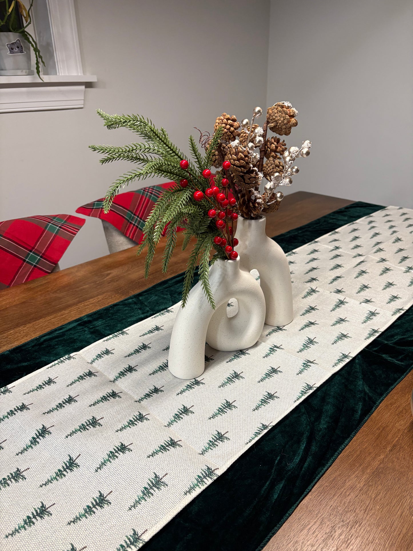 Decorative table setting with a vase of flowers, greenery, and pinecones on a tablecloth with a Christmas tree pattern.