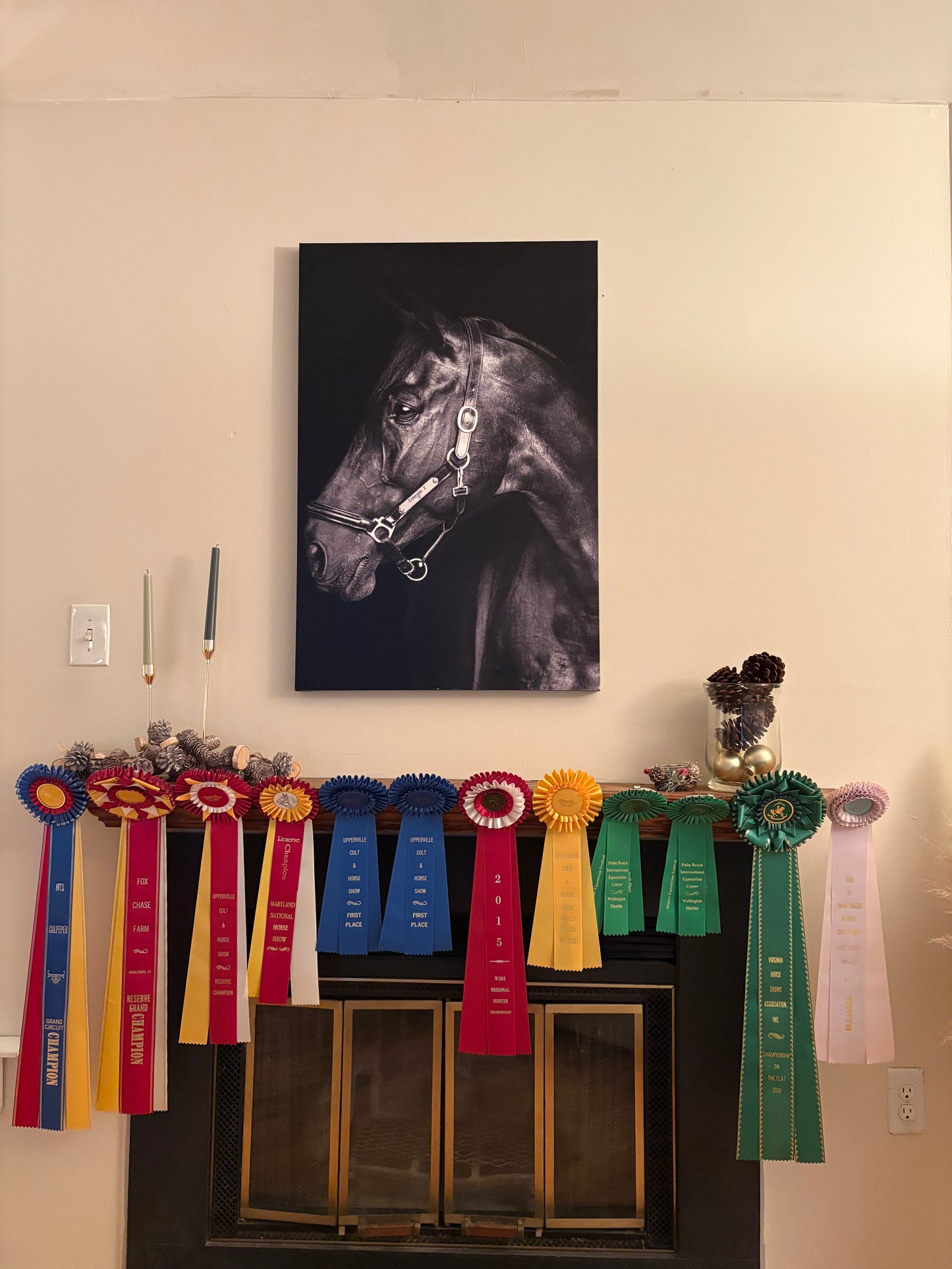 Decorative mantel with ribbons and a framed horse portrait on a wall with pinecones and gold ornaments in a glass jar, candlesticks, and pinecone garland
