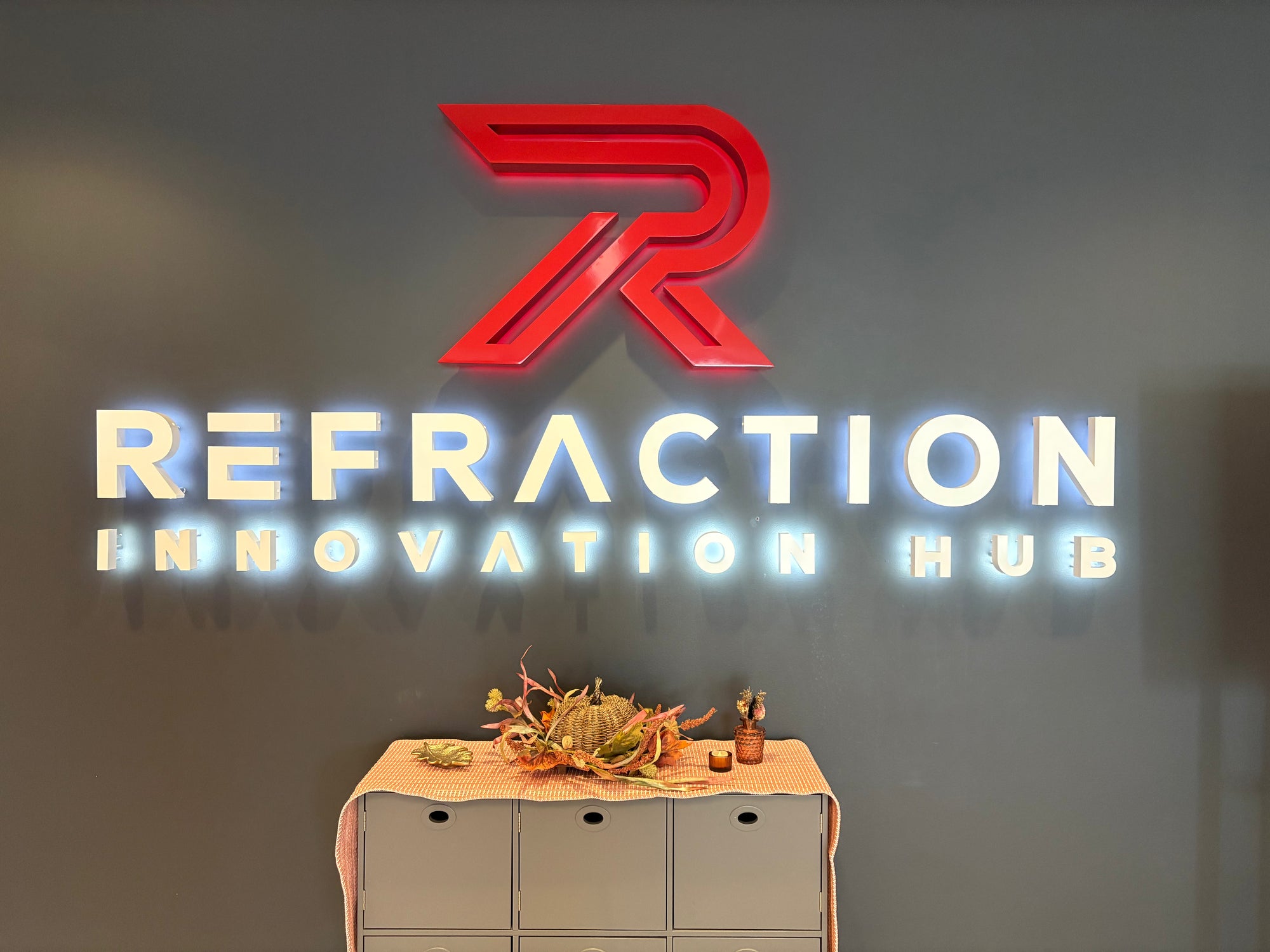 Logo of Refraction Innovation Hub with a decorative table with fall items including a gold leaf tray, woven pumpkin in fall florals, and bud vase in front on a gray background