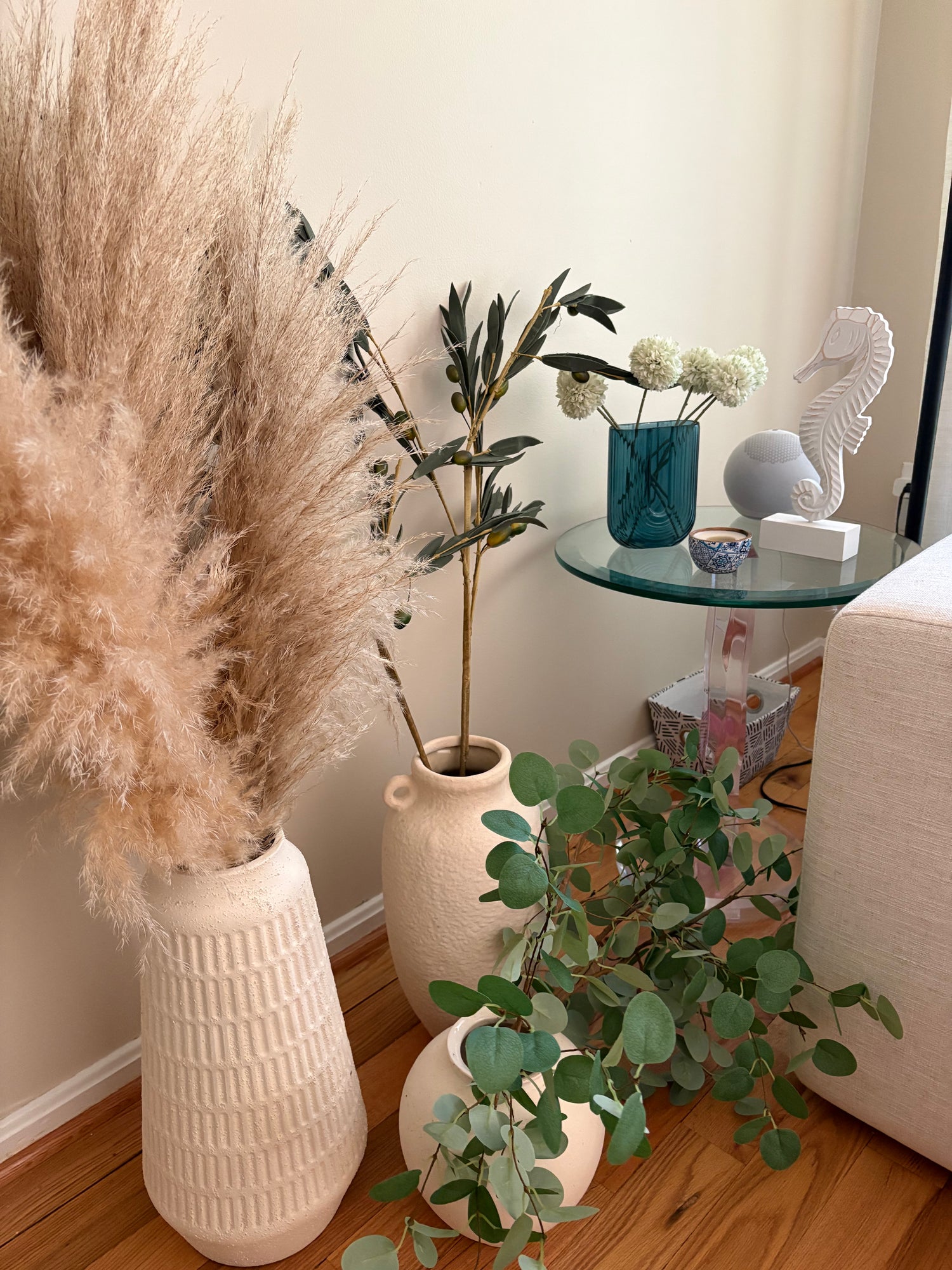 Decorative indoor setting with rental decor including pampas grass, faux olive stems and eucalyptus, and a a blue vase with faux flowers and a white sea horse statue on a glass table.