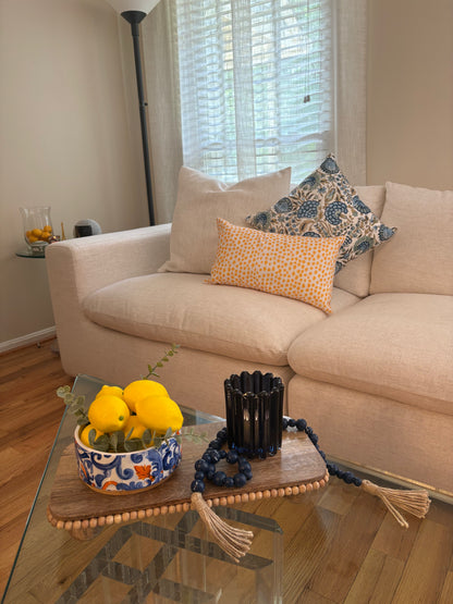 Beige sofa with blue and yellow decorative pillows and a coffee table with summer rental decor including a wooden tray with a bowl full of lemons, a blue beaded tassel  garland, and a blue glass candle holder.