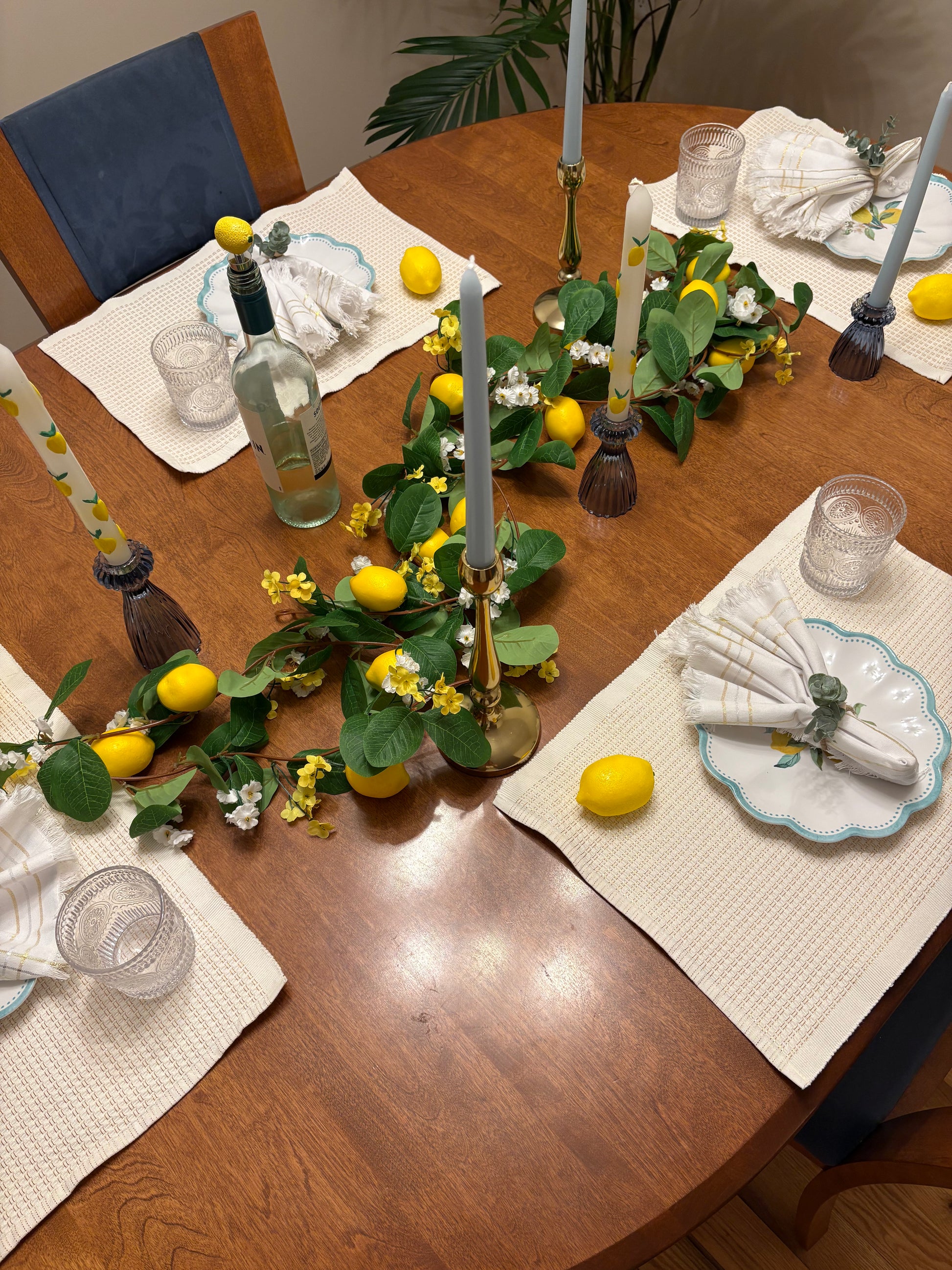 Dining table set with summer rental decor including lemon, candles, garland, candlesticks with tapered tandles, placemats, scalloped plates, and napkins with napkin holders.