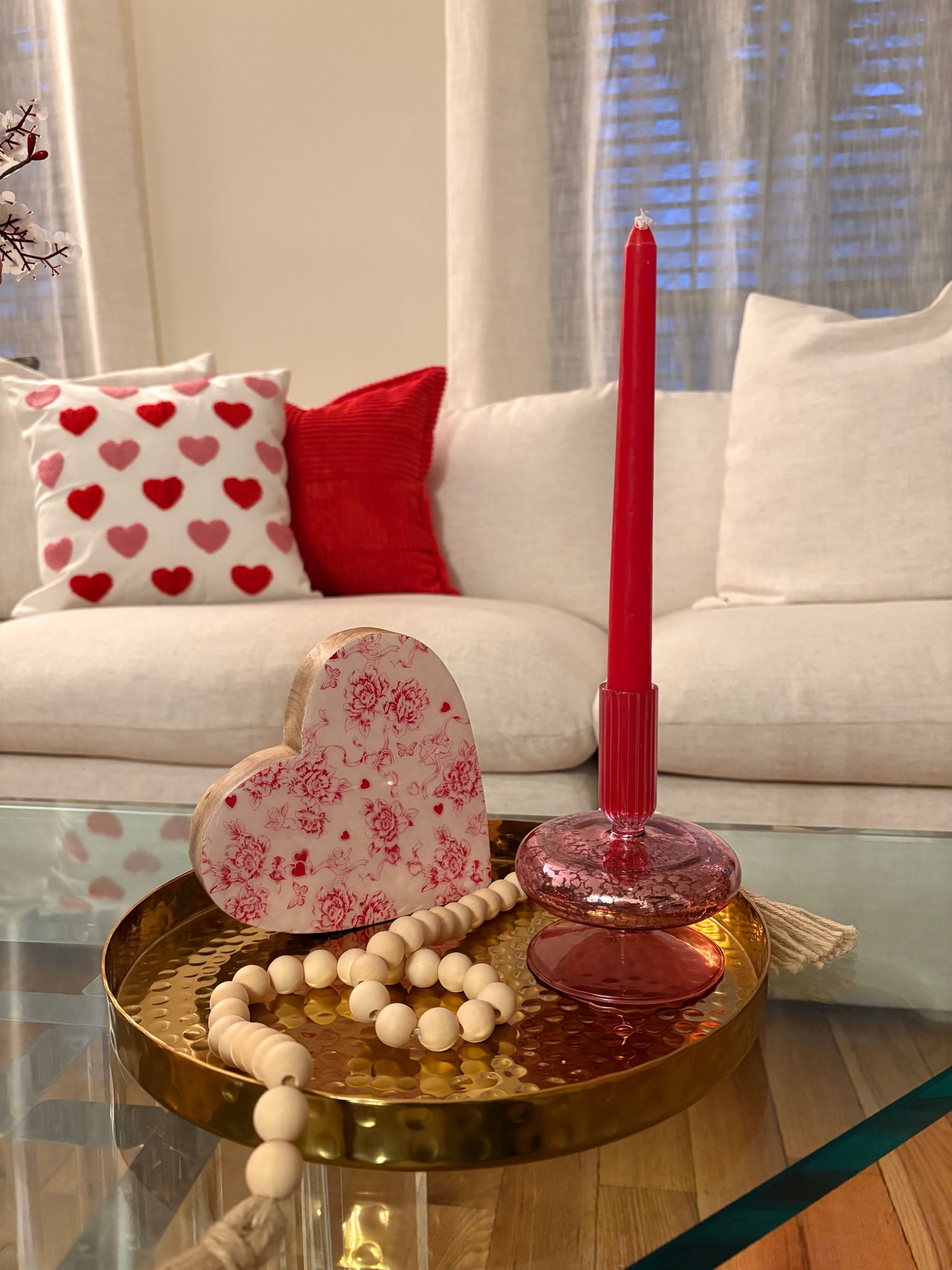 Valentine’s Day rental decor rental styled on a glass coffee table with candle and candlestick, heart with floral pattern, and beaded garland on a gold tray. Beige couch in the background has a pillow with hearts and red pillow.