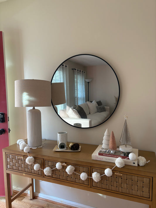 Wooden console table with decorative winter items including Christmas trees, snowflake garland, and candle against a wall with a round mirror.