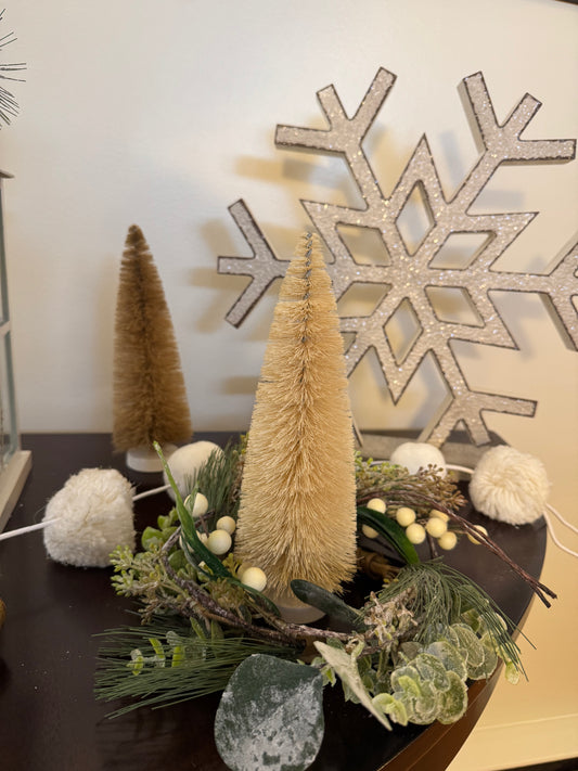 Decorative winter arrangement with beige bottle trees, greenery, and white pom-poms in front of a large snowflake decoration.
