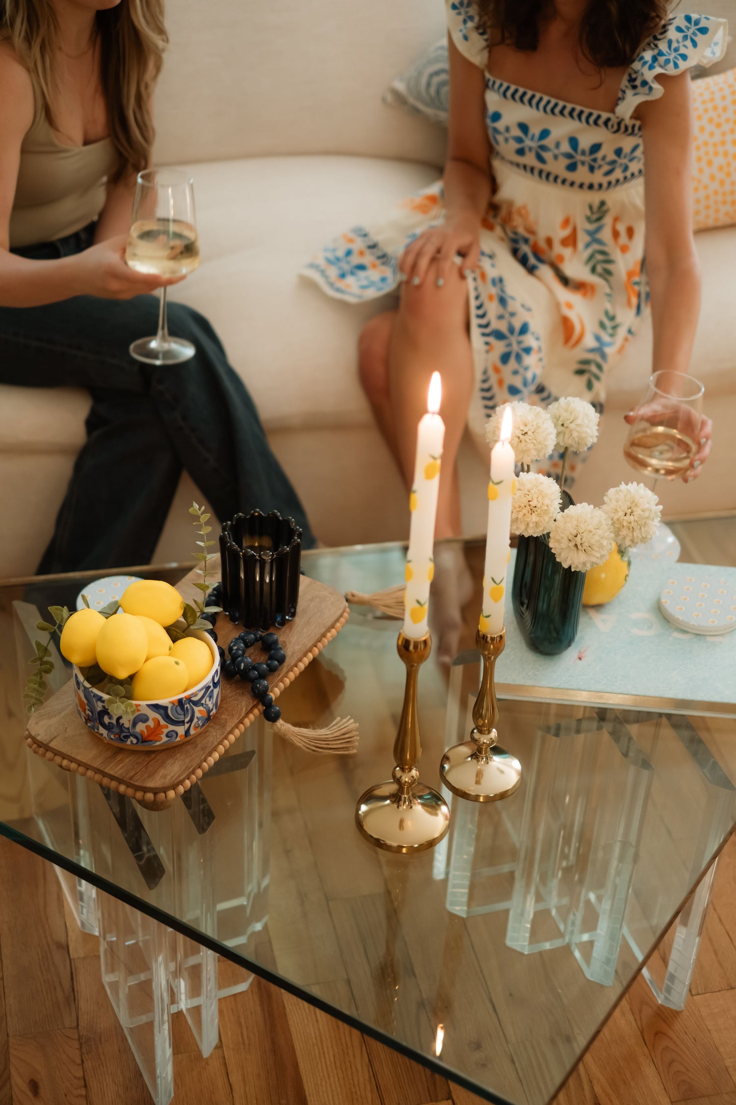 Two women sitting on a couch with a glass of wine and summer rental decor including candles, and lemons on a table.